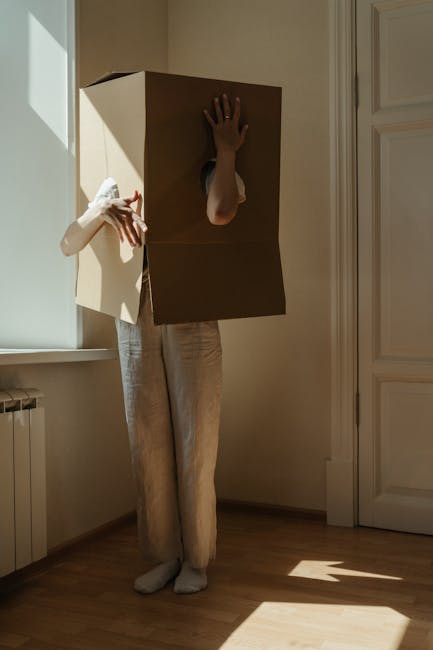 A person dressed in beige trousers and white gloves, with their upper body concealed inside a large cardboard box with a circular hole cut out for the head, stands in a well-lit room next to a window.