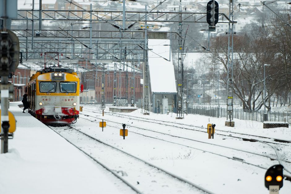 A yellow and red electric train, operated by KRS, is approaching a snow-covered railway station platform in winter, with overhead electrical wires and supporting structures visible above the tracks. T